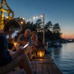 Ung person på brygga i skärgården under midsommarkvällen chattar med vänner på mobilen.